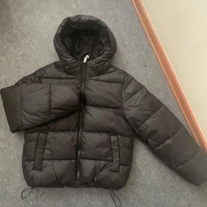 Svart pufferjacka från Gina Tricot - Säljer en svart pufferjacka från Gina Tricot. Jackan har en dragkedja framtill och en mysig huva. Perfekt för kyliga dagar med sin quiltade design och justerbara dragsko i nederkant. Storlek xs. Ny med prislapp