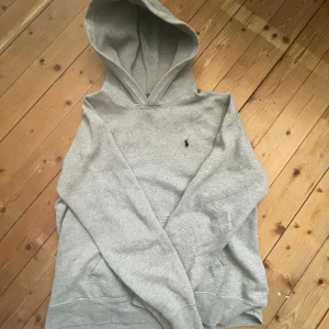Grå hoodie från Ralph Lauren - Säljer en klassisk grå hoodie från Ralph Lauren med en liten broderad logga på bröstet. Tröjan har en bekväm huva och en känguruficka framtill. Perfekt för en avslappnad stil.