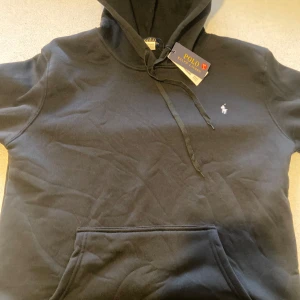 Svart hoodie från Ralph Lauren - Säljer en stilren svart hoodie från Ralph Lauren med klassisk logga på bröstet. Tröjan har en bekväm passform och en praktisk magficka. Perfekt för en avslappnad look.