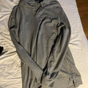 Grå pikétröja från Ralph Lauren - Säljer en stilren grå pikétröja från Ralph Lauren med klassisk krage och knappar. Tröjan har långa ärmar och en liten broderad logotyp på bröstet. Perfekt för en avslappnad men ändå snygg look.