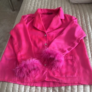 Rosa pyjamas med fjäderdetaljer - Säljer en fantastisk rosa pyjamas med fjäderdetaljer på ärmar och byxben från Nadine Merabi, nypris 1700 kr.  Setet har glittriga knappar och en knytning i midjan. Säljes pga viktnedgång. 