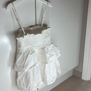 Vit klänning med volanger från Gina Tricot - Söt vit klänning från Gina Tricot med smala axelband och volanger. Klänningen har ett elastiskt livstycke och små guldiga detaljer som ger en glittrig effekt. Perfekt för sommarens varma dagar.