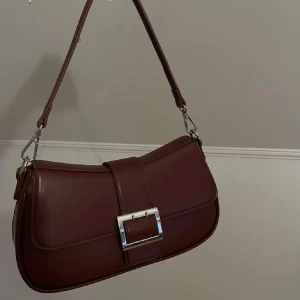 Vinröd axelväska med spänne - Snygg vinröd axelväska i skinn med ett stilrent spänne framtill. Väskan har en justerbar axelrem och en elegant design som passar perfekt för olika tillfällen. Ifrån Åhléns, köpt på Plick men var inte riktigt min still 