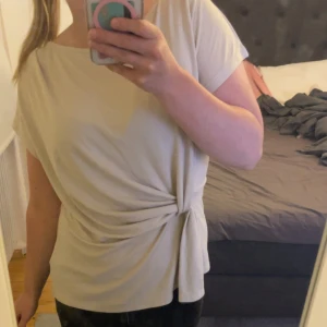 Beige omlottopp med knyt i sidan - Snygg beige topp med omlottdetalj och knyt i sidan. Modellen är kortärmad och har en mjuk, rundad halsringning. Materialet känns lätt och skönt mot huden, perfekt för dig som gillar stilrena och enkla plagg med en twist. Köpt för 600kr 