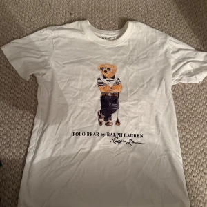 Vit Polo Bear t-shirt från Ralph Lauren - Vit t-shirt från Ralph Lauren med ikoniskt Polo Bear-tryck på bröstet. Björnen har coola solglasögon och randig tröja, vilket ger en chill vibe. Klassisk rund hals och korta ärmar. Perfekt för dig som gillar stilrena och trendiga plagg.
