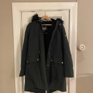 Svart parkas från Barbour med fuskpäls - Svart parkas från Barbour International med huva och fuskpäls. Jackan har guldiga knappar, dragkedja och stora fickor framtill. Insidan är fodrad med mjukt material och quiltad för extra värme. Perfekt för kalla dagar och snygg streetstil.