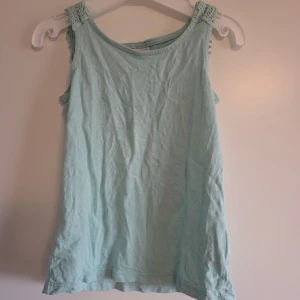 Ljusblå ärmlös linnetopp med spets i ryggen  - Supersöt ljusblå/turkos linnetopp från Lindex med breda axelband och en riktigt snygg virkad spetsdetalj över ryggen. Toppen är ärmlös och gjord i mjuk bomull, perfekt för varma dagar eller när du vill ha något extra fint.