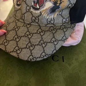 Gucci keps med tiger och GG-mönster - Snygg keps från Gucci med klassiskt GG-monogram i beige och svart, prydd med två broderade tigrar framtill. Kepsen har svart mesh på sidorna och justerbar passform. Perfekt statement-piece för dig som vill sticka ut.