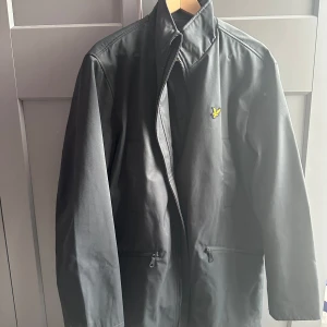 Svart vindjacka från Lyle & Scott - Oanvänd. Skickar iväg samma dag eller dagen efter🙌🏼 Orginal pris är 2099kr. Snygg svart vindjacka från Lyle & Scott med klassisk gul logga på bröstet. Jackan har hög krage, dragkedja framtill och två fickor med dragkedja. Perfekt för dig som vill ha en stilren och enkel jacka med sportig vibe. 