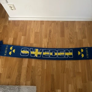 Blå-gul Sweden halsduk med kronor - Snygg blå halsduk med texten SWEDEN i gult och vitt samt tre kronor på sidorna. Perfekt accessoar för att visa ditt stöd för Sverige. Halsduken har fransar i ändarna och är riktigt mjuk och skön.
