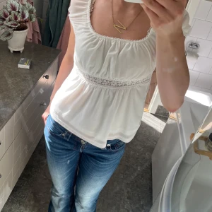 Vit blus med spetsdetalj - Supersöt vit blus med korta ärmar och fyrkantig ringning. Blusen har en romantisk spetsdetalj under bysten och små volanger längs kanterna. Lätt och luftig modell som ger en feminin vibe, perfekt till jeans eller kjol.