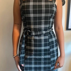 Svart rutig kortklänning med bälte - Snygg svart klänning med vita rutor och breda axelband. Modellen är kort och har ett matchande tygbälte i midjan som knyts med metallring. Passformen är normal och klänningen har en stilren, modern look som funkar till många tillfällen.