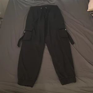 Svarta cargopants med snörning - Svarta cargopants med elastisk midja och snörning. Byxorna har stora fickor på sidorna med lock och banddetaljer, samt resår vid bensluten för en avslappnad look. Perfekta för en streetwear-stil och riktigt sköna att bära.