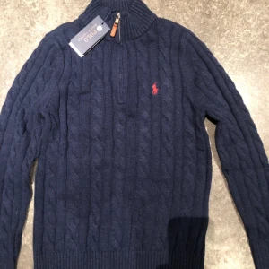 Marinblå stickad tröja Polo Ralph Lauren - Kabelstickad marinblå tröja från Polo Ralph Lauren med halv dragkedja och hög krage. Tröjan har lång ärm, ribbade muddar och röd broderad logga på bröstet. Perfekt för dig som gillar klassisk och stilren look. Den är helt ny och 10/10 skick. Pris i butiker 2000-2500 mitt pris 899