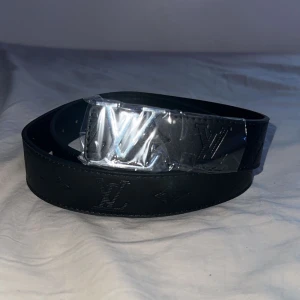 Svart skärp i skinn - Säljer ett svart bälte i skinn med klassiska loggor och monogram präglade längs hela bältet. Bältet har en silverfärgad -spänne. Perfekt accessoar för att lyfta din outfit.