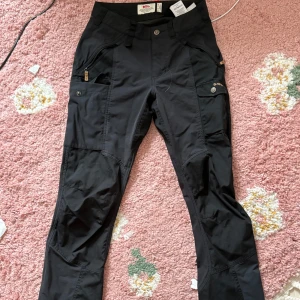 Svarta G-1000 cargopants från Fjällräven - Svarta cargopants från Fjällräven i slitstarkt G-1000-material. Byxorna har flera fickor med dragkedja och knapp, förstärkta knän och klassisk cargomodell. Perfekta för äventyr eller chill utomhus. Snygga detaljer och logga på benet.