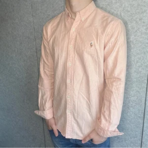 Ljusrosa skjorta Polo Ralph Lauren M - Snygg ljusrosa skjorta från Polo Ralph Lauren i slim fit-modell. Skjortan har klassisk button down-krage, broderad logga på bröstet och knäppning framtill. Tillverkad i bomull och har långa ärmar. Perfekt för en clean och stilren look.