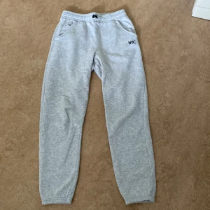 Grå mjukisbyxor med NYC-tryck - Snygga grå mjukisbyxor med elastisk midja och muddar vid bensluten. Byxorna har två fickor och ett litet svart NYC-tryck på ena sidan. Perfekta för chill dagar eller när du vill vara bekväm. Tillverkade i mjukt bomullsmaterial.