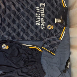Real Madrid fotbollströja set blå - Snygg Real Madrid fotbollströja och shorts i mörkblått med gula och vita detaljer. Tröjan har ett vågigt mönster, klubbmärke på bröstet och sponsortryck. Tillverkad i lätt och ventilerande material, perfekt för träning eller match.