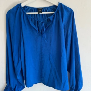 Blå blus med knyt från Lindex XS - Blus i klarblå färg från Lindex, storlek XS. Modellen har långa ärmar med resår vid ärmslut och en snygg v-ringning med knyt framtill. Luftig och skön passform