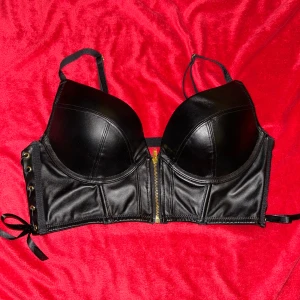 Svart korsett med snörning och dragkedja - Snygg svart korsett med push-up kupor och justerbara axelband. Framsidan har en guldfärgad dragkedja och sidorna är dekorerade med snörning och svarta rosetter. Materialet är glansigt och ger en cool vibe, perfekt för att sticka ut. Baksidan har elastiska band för extra passform. Från Hunkemöllers Private Collection