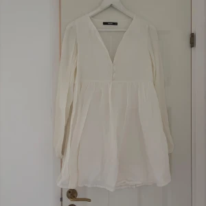 Vit kortklänning med v-ringning - Säljer en söt vit kortklänning från Bik Bok med v-ringad hals och knappar framtill. Klänningen har långa ärmar och ett luftigt, flowy tyg som ger en chill vibe. Perfekt för varma dagar och kan stylas med sneakers eller boots.