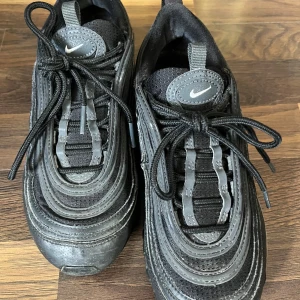 Nike Air Max 97 svarta sneakers - Svarta Nike Air Max 97 med ikoniska vågformade linjer och synlig Air-sula. Ovandel i textil och syntet, med reflekterande detaljer och klassisk snörning. Swoosh-logga på sidan och tungan. Perfekta för dig som gillar streetstyle och sportig vibe.