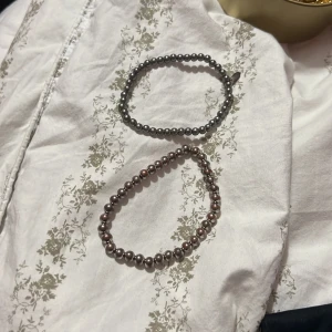 Två pärlarmband i silver och brunt - Säljer två stilrena pärlarmband, ett i metallic silver och ett i mörkbrun ton. Båda har rundade pärlor och elastisk tråd som gör dem enkla att ta på. Perfekt för dig som gillar minimalistiska accessoarer med lite glans.