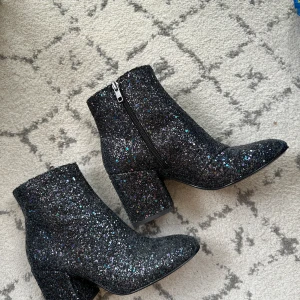 Glittriga svarta stövletter från Ash - Snygga svarta stövletter från Ash täckta i glittrigt material med chunky klack och spetsig tå. Skorna har dragkedja på sidan och massor av glitter i olika färger som ger en riktigt festlig vibe. Perfekta för dig som vill sticka ut med din stil.