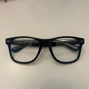 Ray-Ban Wayfarer - Klassiska svarta Wayfarer-glasögon från Ray-Ban med bred båge i blank plast och tydlig logga på skalmarna. Rektangulär form som ger en tidlös och cool vibe. Perfekta för dig som vill ha en ikonisk look med stilren känsla.