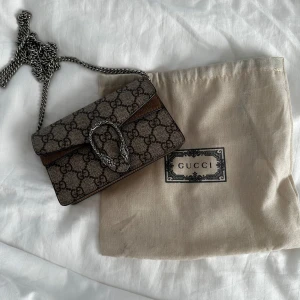 Gucci Dionysus axelväska med kedja - Säljer en Gucci Dionysus axelväska med klassiskt GG-monogram i beige och brunt. Väskan har en silverfärgad kedja och ett snyggt spänne med tigerhuvuden. Utsidan är i canvas och insidan har detaljer i mocka och skinn. Perfekt storlek för det viktigaste.
