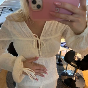 Vit blus med knyt och v-ringning - Säljer en vit blus med v-ringning och snygga knytband framtill samt vid ärmsluten. Blusen har ett lätt rynkat tyg och långa ärmar med voluminösa muddar