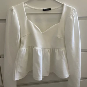 Vit blus med volang från Missay - Supersöt vit blus från Missay med lång ärm och v-ringad hals. Blusen har en volang nedtill som ger en extra touch och är tillverkad i mjukt bomullsmaterial. Perfekt för dig som gillar en feminin och trendig look.