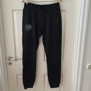 Svarta mjukisbyxor Hammarby - Svarta mjukisbyxor från Craft med dragsko i midjan och ribbade muddar vid bensluten. Byxorna har en diskret logga på ena benet och är tillverkade i ett mjukt och bekvämt material som passar perfekt för träning eller chill.