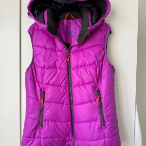 Lila dunväst med huva från H&M - Snygg lila dunväst från H&M med huva och svarta detaljer. Västen har två fickor med dragkedja och orange zip-pullers. Perfekt för lager-på-lager och chillare dagar när du vill hålla värmen utan att bli för varm.