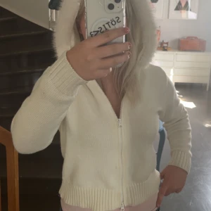 Vit hoodie med fluffig huva - Supermysig vit hoodie med fluffig pälsdetalj på huvan. Tröjan är stickad och har dragkedja framtill samt ribbade muddar vid ärmslut och nederkant. Perfekt för kyliga dagar när du vill vara både varm och snygg. Passar dig som gillar en chill och cozy stil.