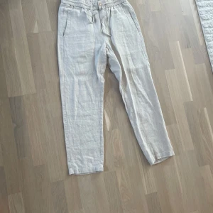 Beiga linnebyxor från Skogen, S - Snygga och luftiga beiga linnebyxor från Skogen i 100% linne. Byxorna har elastisk midja med snörning och raka ben, perfekta för en chill och avslappnad stil. Fickor på sidorna och en enkel knapp framtill.