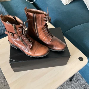 Bruna skinnkängor med snörning och dragkedja - Snygga bruna kängor i skinn med snörning framtill, dragkedja på sidan och coolt spänne vid ankeln. De har platt sula och rund tå, perfekt för dig som gillar en klassisk men ändå edgy stil. Passar till både jeans och kjol för en chill look.