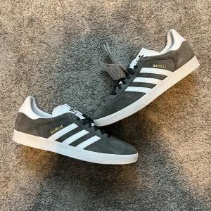 Adidas Gazelle grå/vita sneakers mocka - Adidas Gazelle sneakers i grå mocka med vita detaljer och klassiska tre ränder på sidorna. Skorna har svart snörning, vit plös och hälkappa med Adidas-logga. Platt vit sula med mönster undertill. Perfekta för en clean och stilren look. Str 45 box tillkommer samt vita skosnören som alternativ. Pris kan diskuteras 