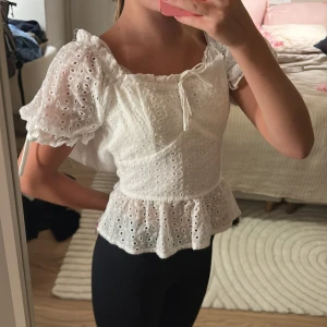 Vit blus med volang från Meet Me There - Supersöt vit blus med broderade hål, puffiga korta ärmar och volang nertill. Söt knytning vid halsen och romantisk känsla. Perfekt för dig som gillar en feminin och trendig stil.