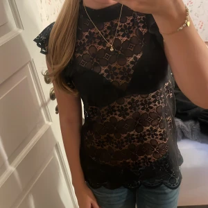 Svart spetsblus från JDY - Supersnygg svart blus från JDY i spets med blommigt mönster. Modellen är ärmlös med volangdetaljer på axlarna och har en hög hals. Perfekt för dig som gillar en feminin och trendig look. Blusen är genombruten och har fina vågiga kanter nertill.Säljer pga att den aldrig kommit till användning💞