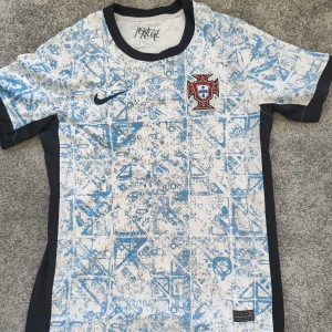 Portugal Nike fotbollströja blåvit storlek S - Säljer en Portugal fotbollströja från Nike med unikt blått grafiskt mönster på vit bakgrund. Tröjan har svarta detaljer vid krage och ärmslut, samt Portugal-emblem broderat på bröstet. Materialet är lätt och andas, perfekt för match eller träning.