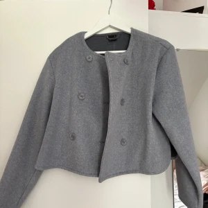 Grå kort kappa med knappar - Stilren grå kappa i kort modell med rund halsringning och dubbelknäppning framtill. Jackan har långa ärmar och är tillverkad i ett mjukt ullmaterial som ger en clean och modern look. Perfekt för dig som gillar minimalistisk stil.