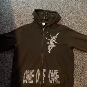 Svart hoodie One of One med strass - Svart hoodie från One of One i storlek M med dragkedja, huva och känguruficka. Framsidan har en stor strassdekorerad ängel och texten 'ONE OF ONE' i glittriga stenar. Perfekt statement-plagg för dig som gillar unika detaljer.