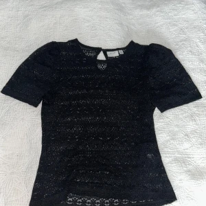 Svart spetsblus från Vila - Supersnygg svart blus från Vila i spets med korta ärmar och rund halsringning. Blusen har ett diskret knappslå bak i nacken och är helspetsad med fina detaljer. Perfekt att styla med jeans eller kjol för en trendig look.