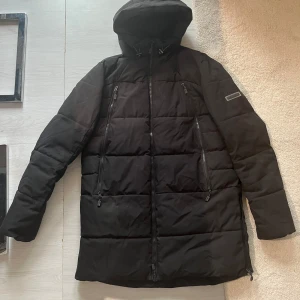 Svart vinterjacka från Kings Will Dream - Svart lång pufferjacka från Kings Will Dream med huva och flera dragkedjor framtill. Jackan har quiltad design och raka linjer, perfekt för kalla dagar. Snygg logga på ärmen och praktiska fickor med dragkedja. Passformen är normal och jackan är fodrad för extra värme.