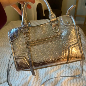 Glittrig silver handväska med dragkedja - Säljer en glittrig silver handväska med både handtag och axelrem. Väskan har en snygg metallic finish, dragkedja upptill och ett extra fack med dragkedja på framsidan. Perfekt för dig som vill sticka ut med en trendig accessoar.