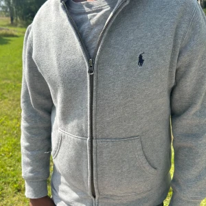Grå hoodie från Polo Ralph Lauren - Säljer en grå hoodie från Polo Ralph Lauren med dragkedja och klassisk logga på bröstet. Tröjan har huva, två fickor fram och är tillverkad i mjuk bomull med fleece på insidan. Perfekt för chill dagar och sköna promenader. Och väldigt ny bara använd ett fåtal gånger