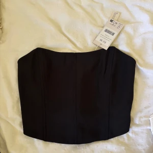 Svart korsett-topp från nakd - Snygg svart korsett-topp från nakd med stilren design och markerade sömmar framtill. Toppen är axelbandslös och har en smockad baksida för extra komfort. Perfekt att styla med höga jeans eller kjol för en trendig look.