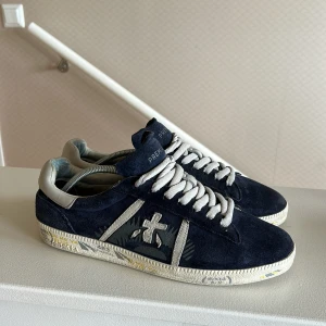 Mörkblå sneakers från Premiata mocka - Snygga mörkblå sneakers från Premiata i mjuk mocka med vita detaljer och snörning. Skorna har en vit sula med tryck och grå logga på sidan. Perfekta för dig som gillar stilrena och unika sneakers med coola detaljer.